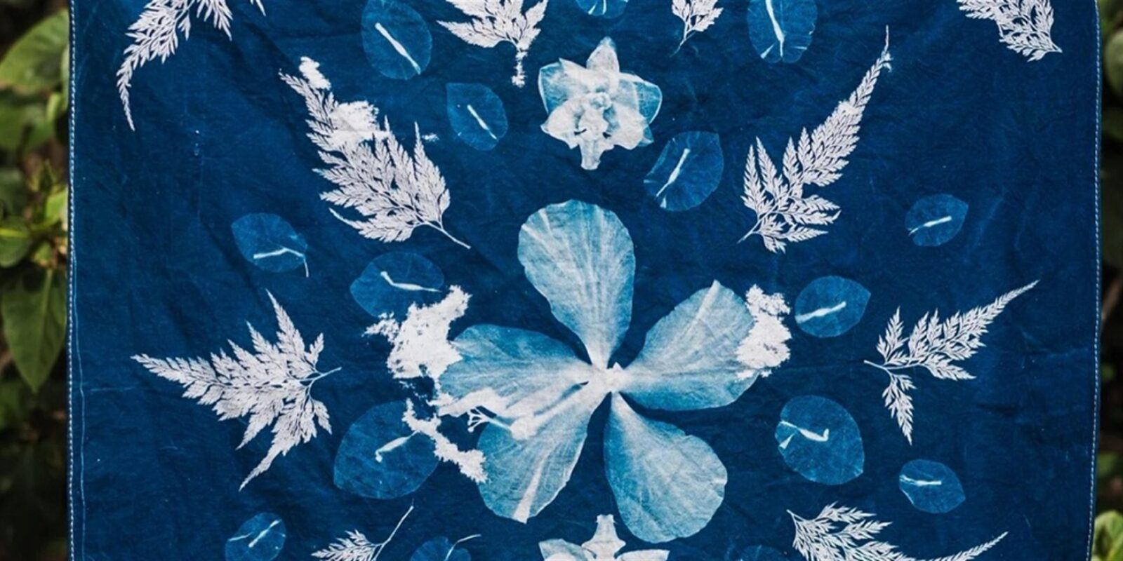 A deep blue cyanotype bandana hung outdoors, printed with white silhouettes of ferns, leaves, and a large flower.