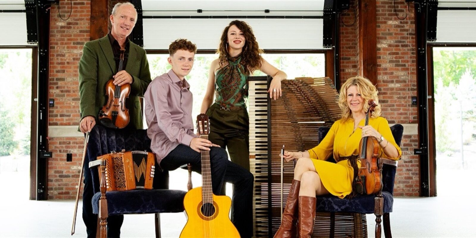 Four musicians in a brick-walled hall with their instruments — two fiddlers, a young man with a guitar, and a young woman beside a piano — the family Celtic ensemble.
