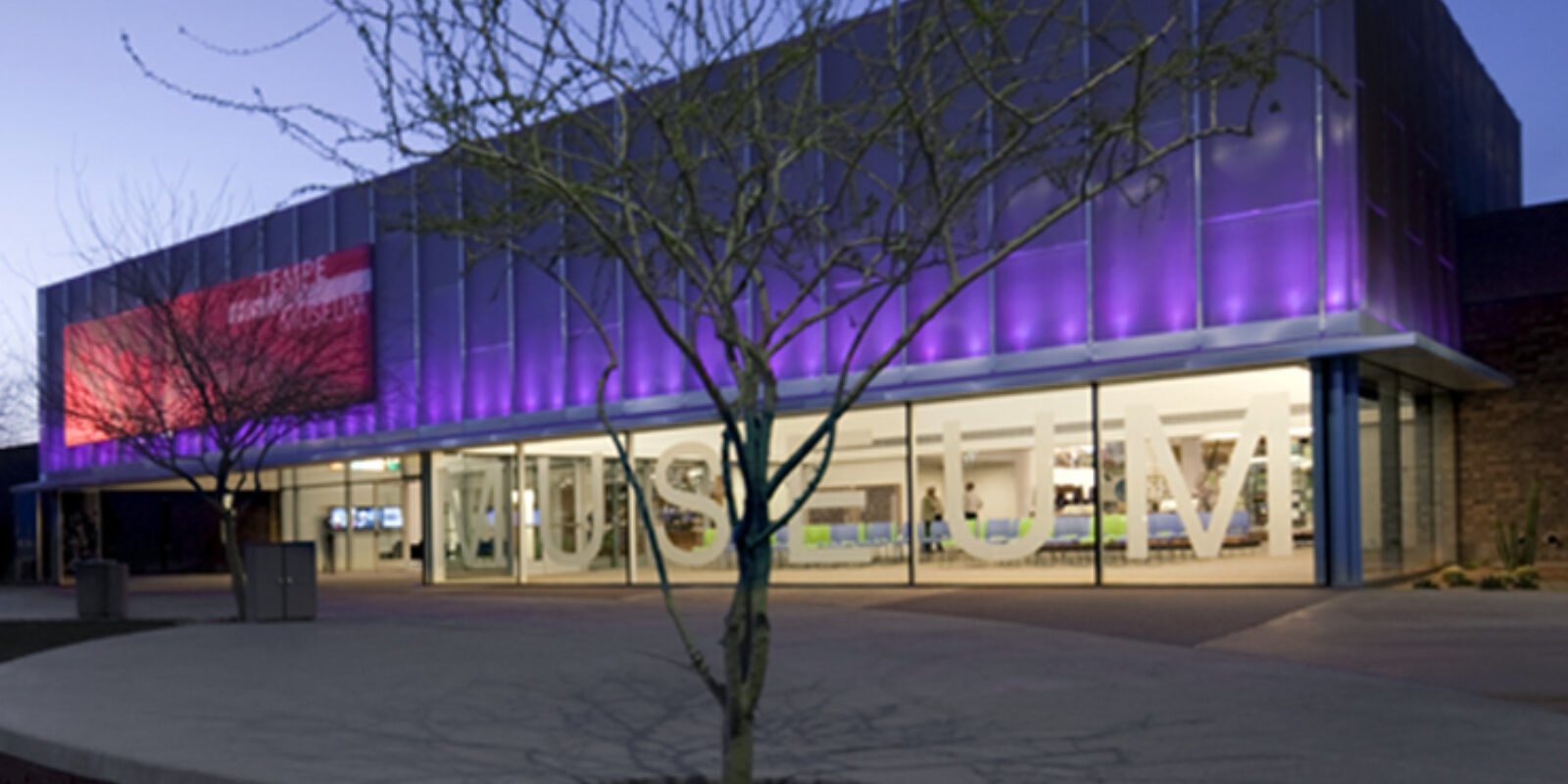 Tempe History Museum at dusk, its modern glass facade lit with purple uplighting and large MUSEUM letters visible through the floor-to-ceiling windows.