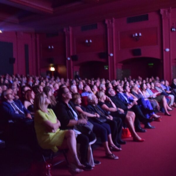 A full audience seated in a red-walled theater, watching the stage intently.