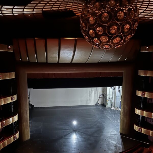 An empty theater auditorium seen from the balcony, its tiers and chandelier in shadow, lit only by a single ghost light standing on the bare stage.