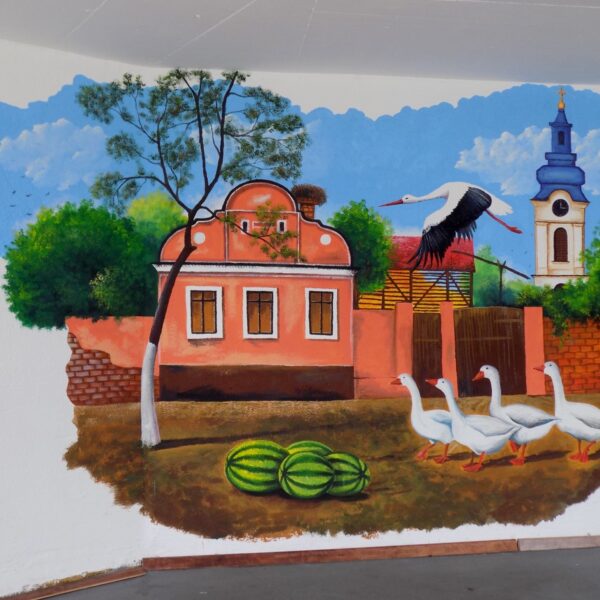 A painted wall mural of a village scene: a pink house with a stork nesting on the chimney, geese in the yard, and watermelons on the ground.