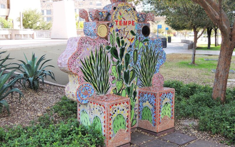 A mosaic-tiled 'Greetings from Tempe AZ' sculpture decorated with cactus and hummingbird motifs, set in a shaded park.